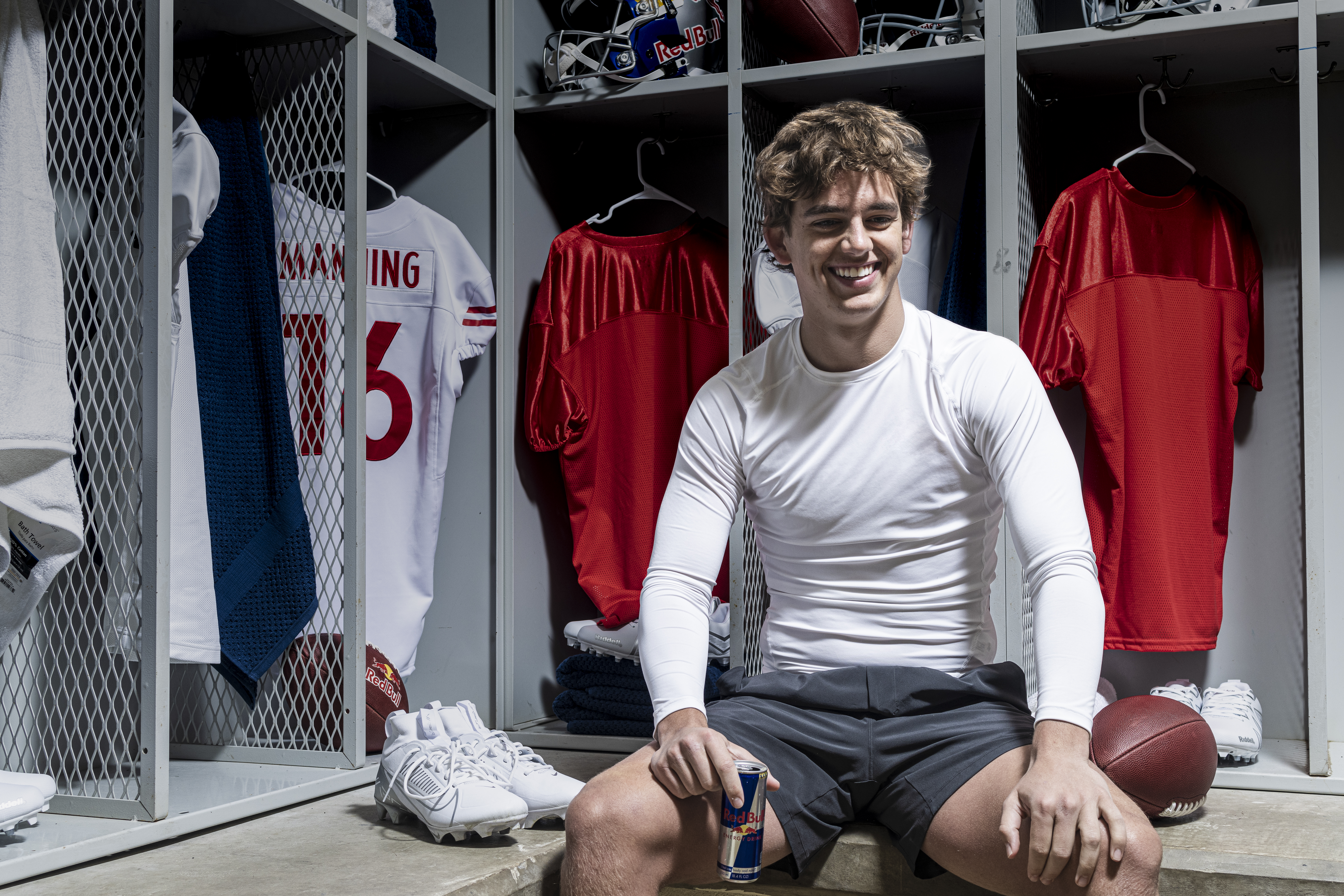 Arch Manning in the locker room, wearing a white long-sleeve shirt and shorts, holding a Red Bull. Football gear is visible around him.
