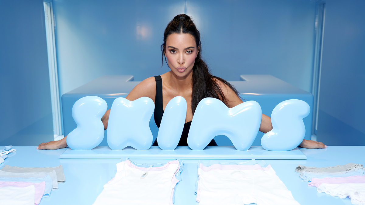 Kim Kardashian poses behind a "SKIMS" sign in a blue room, with clothing items displayed in front of her.