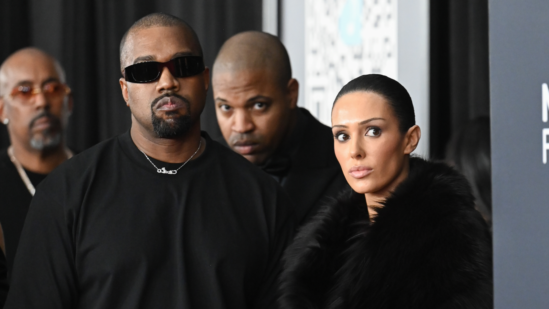Kanye West wearing sunglasses and a black shirt, standing next to Bianca Censori in a black fur coat, with two men in the background.