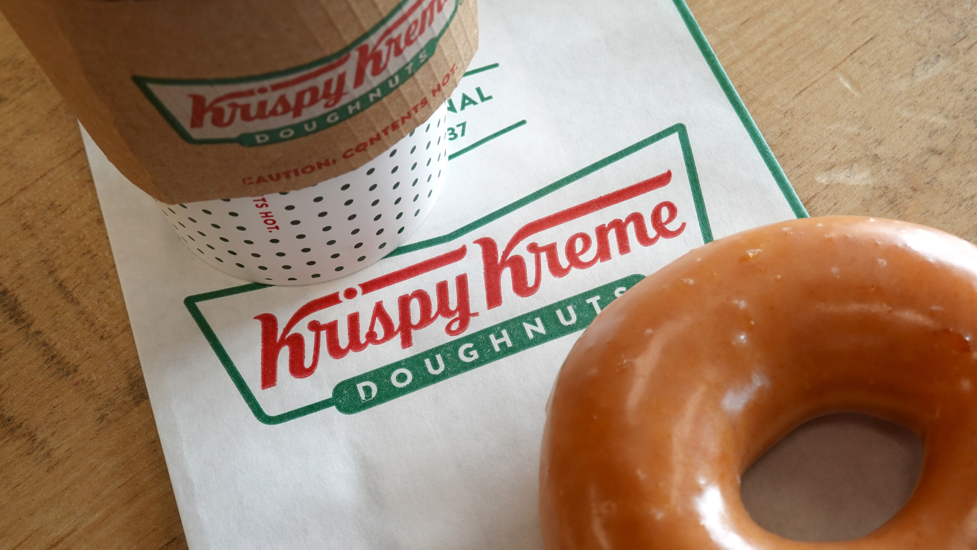 Krispy Kreme coffee cup and glazed doughnut on a branded napkin on a wooden table.